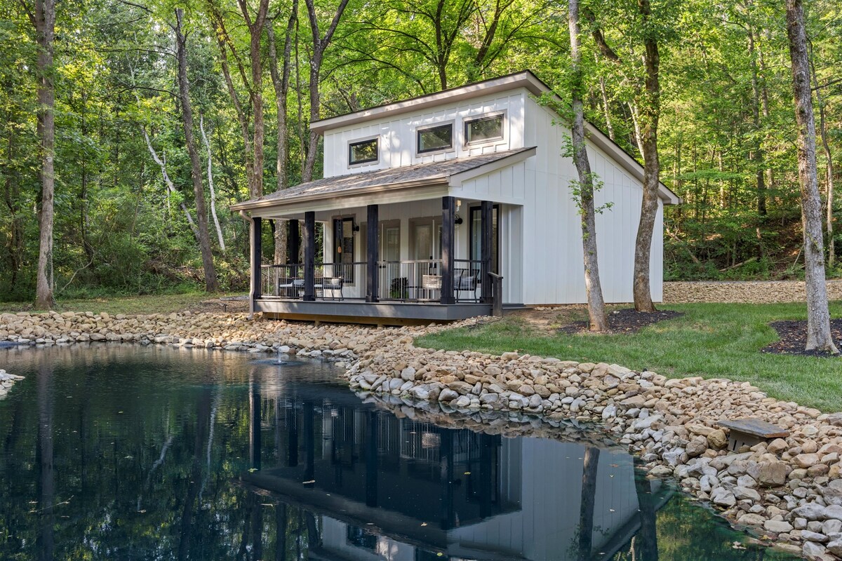 A modern white tiny home is set amid lush greenery, overlooking a calm lagoon. The structure features a covered porch with seating for relaxation. Stones encircle the water’s edge, reflecting the surrounding trees and natural beauty.