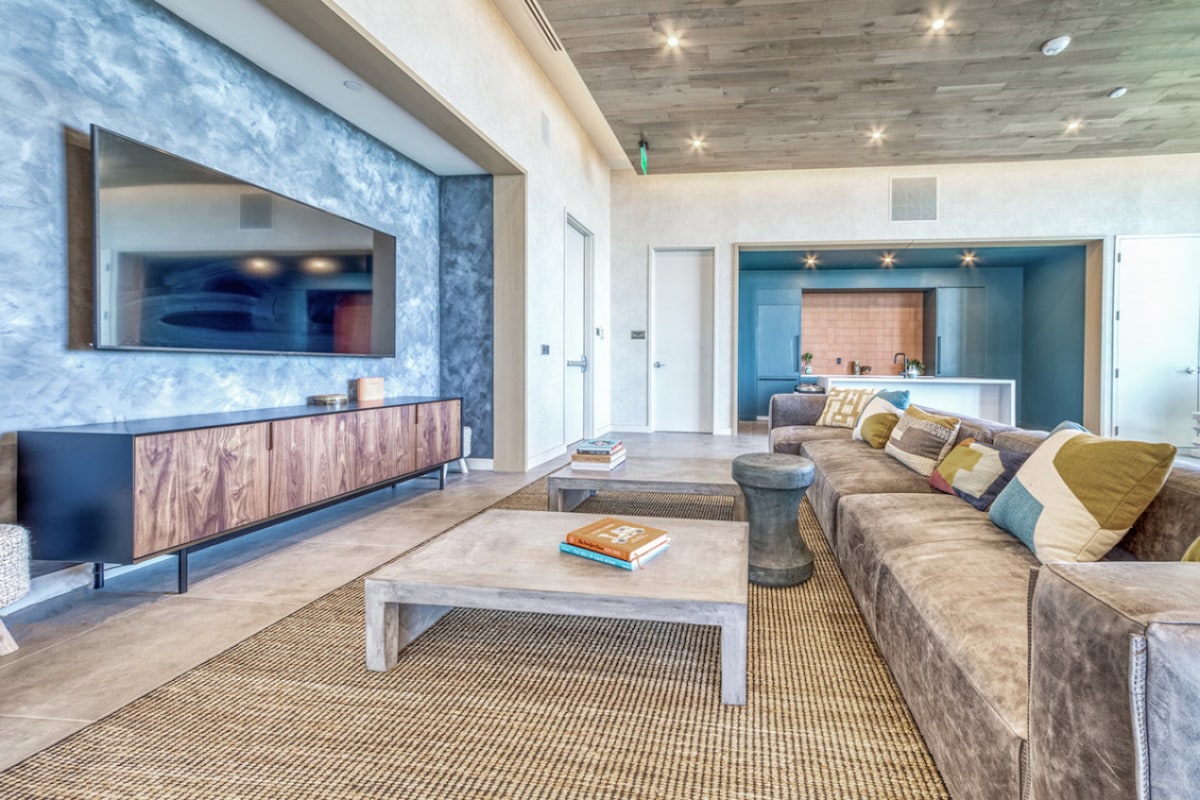 The living area features a sectional sofa adorned with colorful throw pillows, positioned across from a mounted flat-screen TV. A coffee table made of light wood sits atop a textured area rug. Soft lighting highlights the contemporary decor and open kitchen space in the background.
