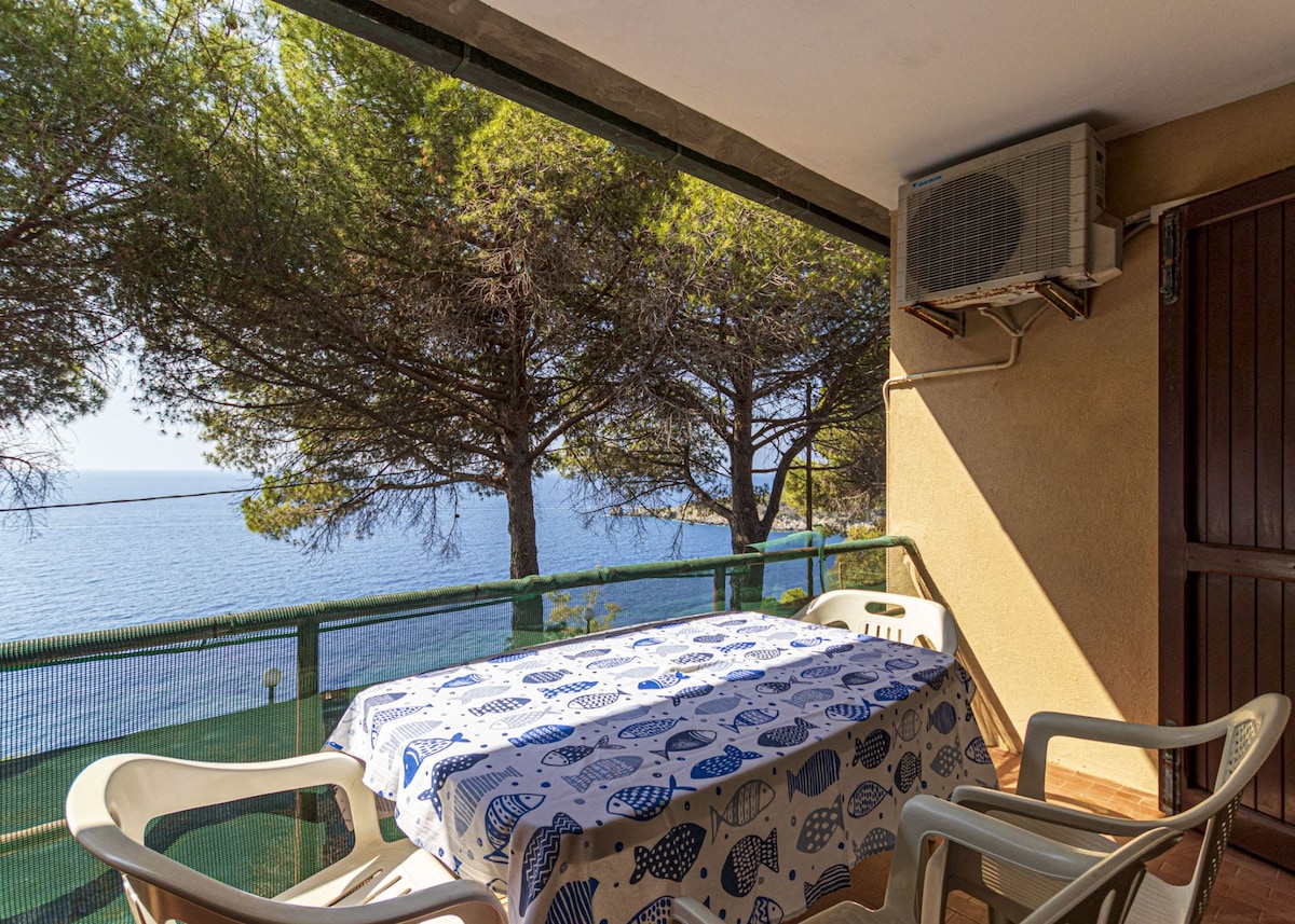 An outdoor terrace overlooks the sea, featuring a table with a patterned tablecloth and several white chairs. Tall trees provide shade, while an air conditioning unit is mounted on the wall, ensuring comfort in warmer weather.