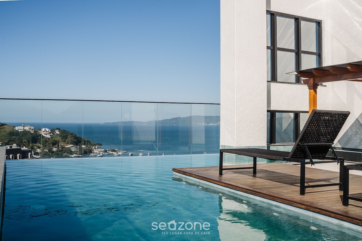 A rooftop pool is visible, featuring clear water that reflects the surrounding landscape. Lounge chairs are positioned beside the pool, offering a view of the ocean and nearby coastline. The area is well-lit by natural sunlight, enhancing the serene atmosphere.