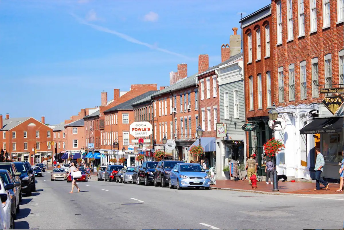 A bustling downtown street is lined with historic brick buildings, featuring a variety of shops and restaurants. Parked vehicles are visible along the curb, and pedestrians are seen enjoying the vibrant atmosphere. Clear blue skies and a warm light enhance the lively scene.