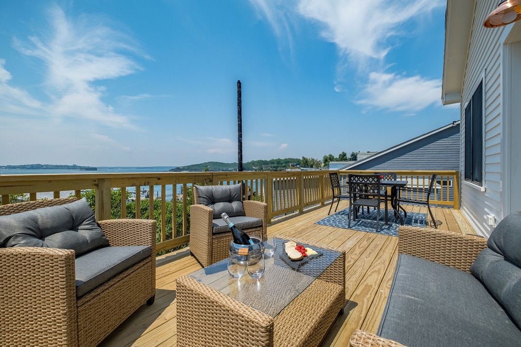 An outdoor deck features a spacious seating area with two woven chairs and a glass-topped table. A dining table with four chairs is positioned to enjoy unobstructed views of the water. Natural light brightens the space, while scenic surroundings create a peaceful atmosphere.
