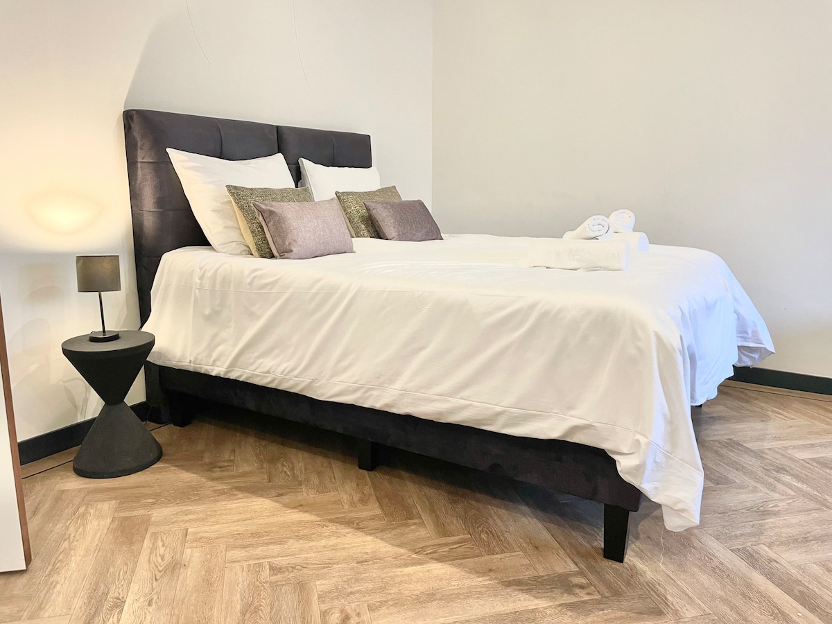 A spacious bed with a dark upholstered headboard is dressed in crisp white linens. Decorative pillows add texture and warmth. A small, modern lamp atop a minimalist side table provides soft lighting, complementing the light-coloured walls and wood-patterned flooring.