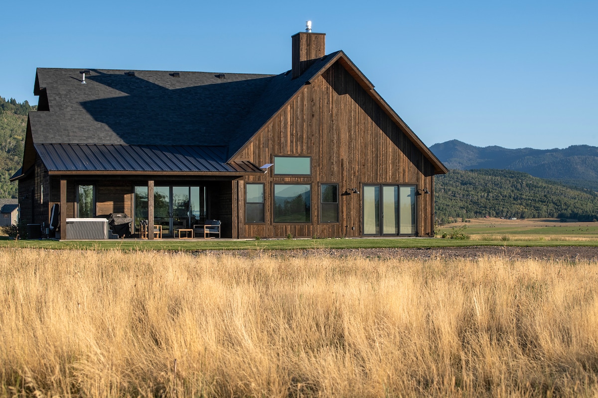 A modern rustic home is depicted with a sloping roof and a combination of wood and metal accents. Large windows allow for ample natural light, showcasing unobstructed views of surrounding grassy fields and distant mountains.