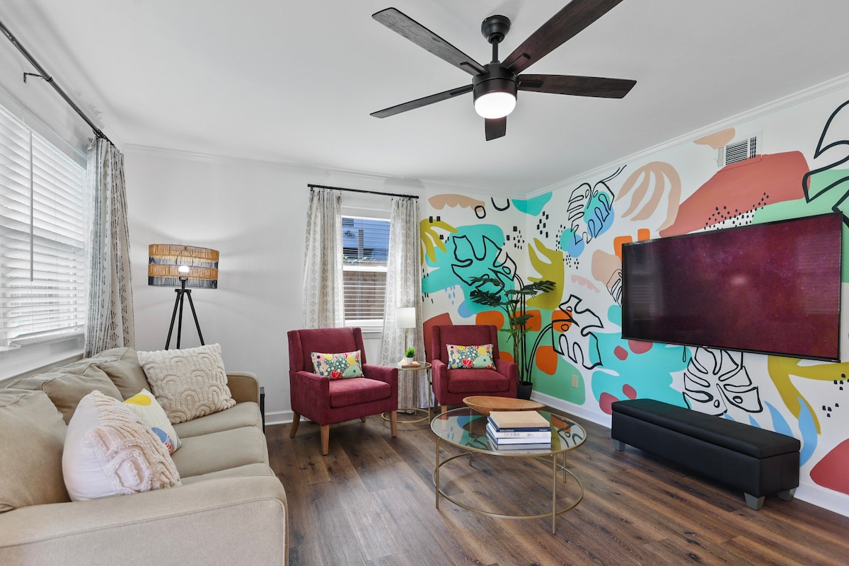 The living room features a relaxing seating arrangement with a light-colored sofa and two red accent chairs. A stylish glass coffee table is positioned in front, while a vibrant mural decorates one wall. Natural light filters through the windows, complemented by a ceiling fan overhead.