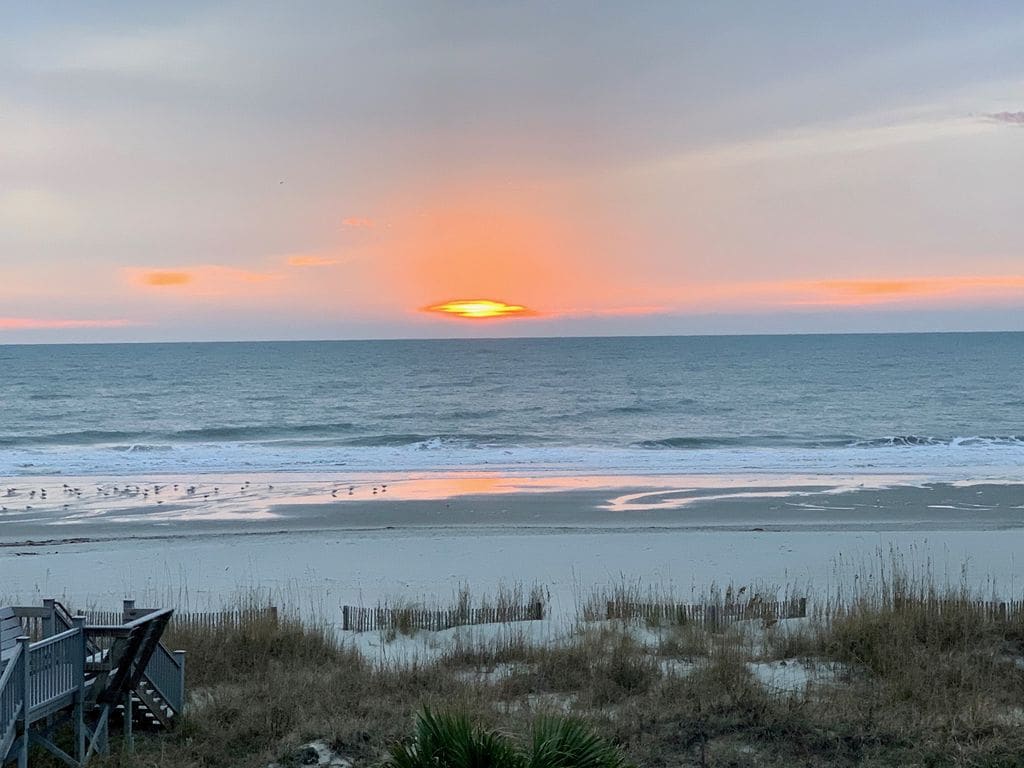 A serene ocean view captures the gradient of colors in the sky during sunset, reflecting on the calm waters. The sandy beach is visible in the foreground, with grasses and beach vegetation framing the scene, inviting a sense of tranquility.