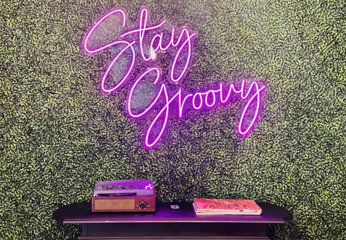 A vibrant neon sign reading 'Stay Groovy' glows in pink against a textured green wall. A retro record player rests on a sleek black table, accompanied by a stack of colorful vinyl records, creating a lively and inviting decorative arrangement.