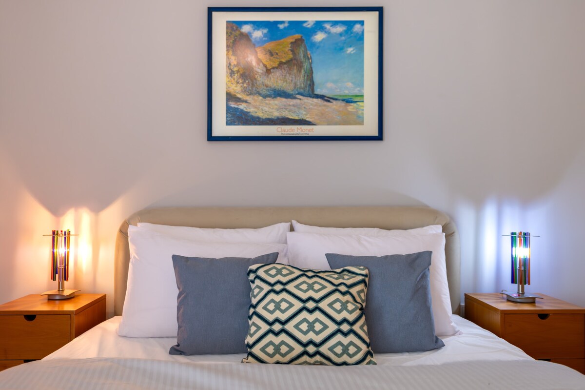 A serene bedroom is depicted, featuring a cozy queen-size bed adorned with multiple pillows. Bedside tables with colorful lamps are positioned on either side, and a framed landscape artwork hangs above the headboard. Soft lighting casts a warm glow in the tranquil space.