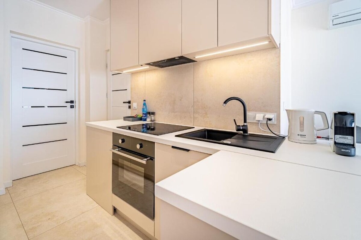 A modern kitchen features sleek cabinetry and a clean countertop. An oven and stovetop are integrated into the design, along with a contemporary sink. Essential appliances, including a kettle and coffee maker, are neatly arranged for convenience.