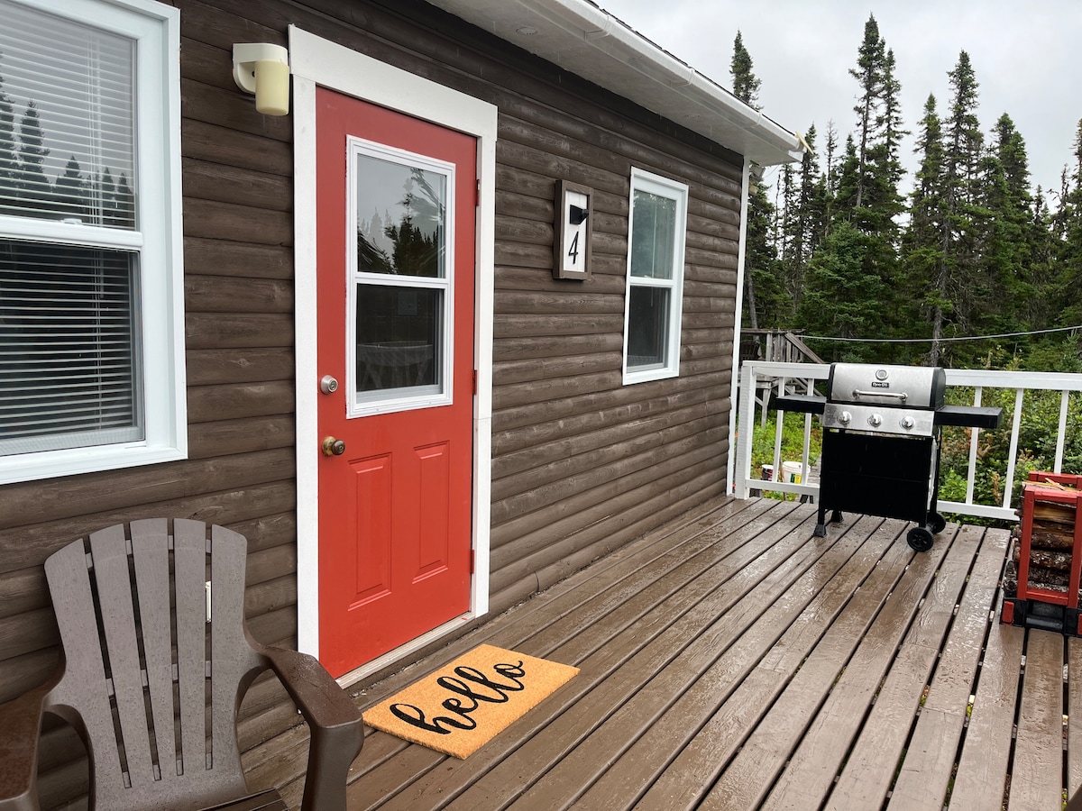 The entrance of Cabin 4 features a vibrant red door, complemented by a welcoming doormat. A wood chair sits on the patio, which is equipped with a propane grill and a small cart with firewood, surrounded by greenery in the background.