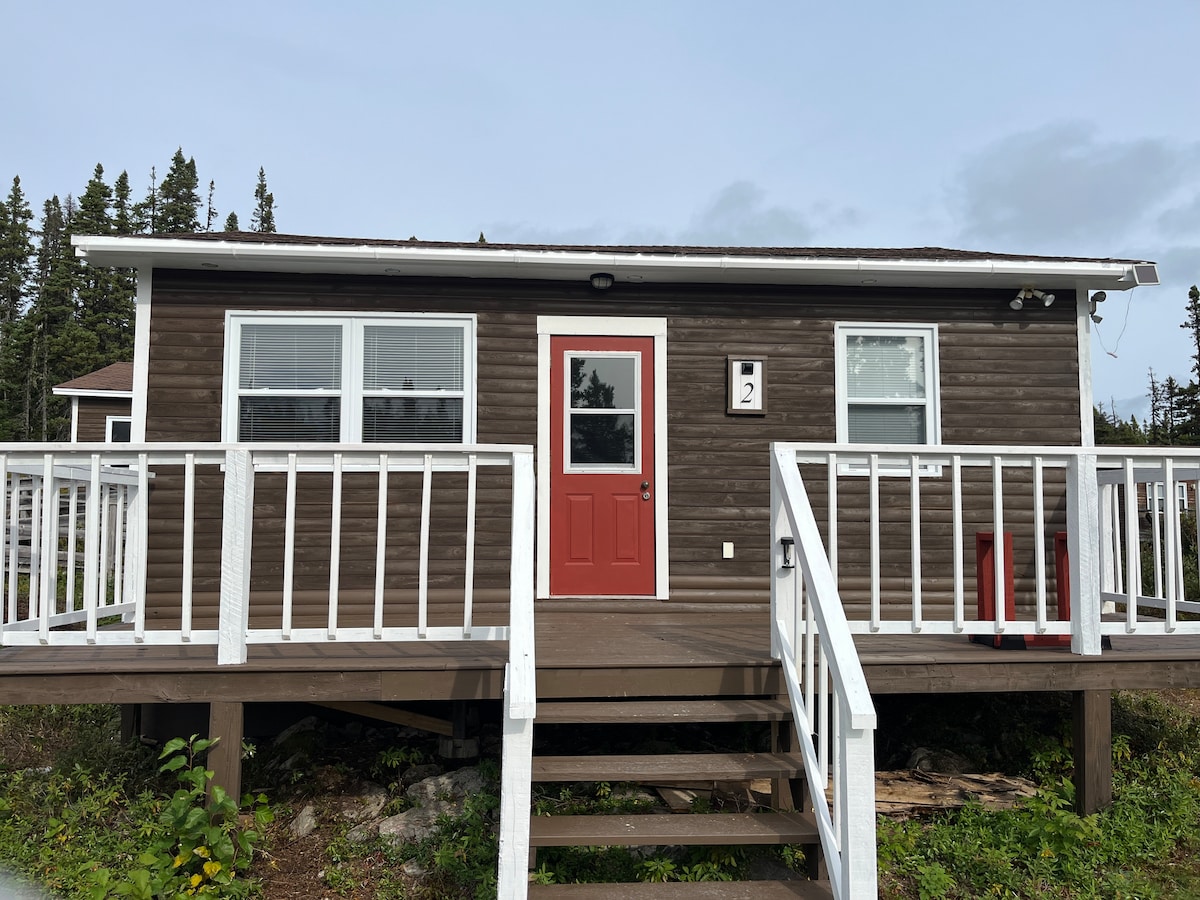 A cabin exterior is featured with a welcoming red front door. White railings line the entrance steps, leading to a single-story structure with two windows on either side of the door. Surrounding greenery adds to the natural setting.