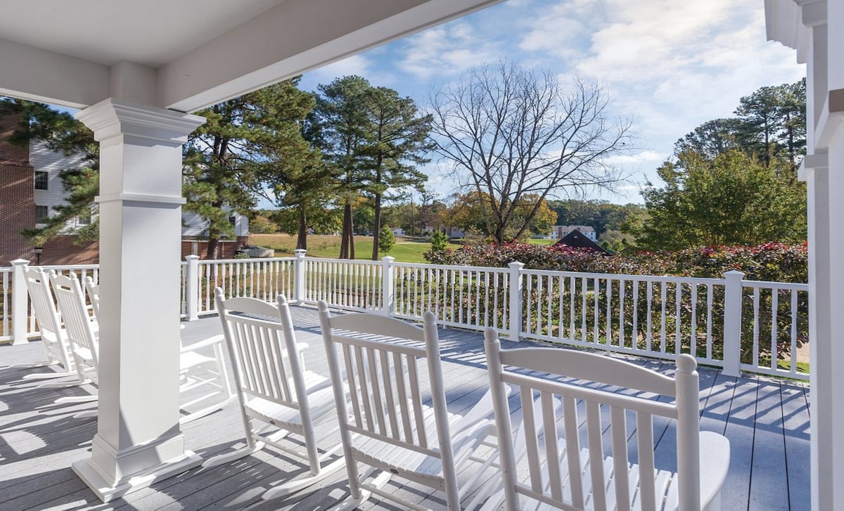 A spacious balcony is furnished with white rocking chairs, inviting relaxation. Lush greenery and trees are visible in the distance, creating a serene outdoor setting. The railing adds safety, while the blue sky with soft clouds enhances the peaceful ambiance.