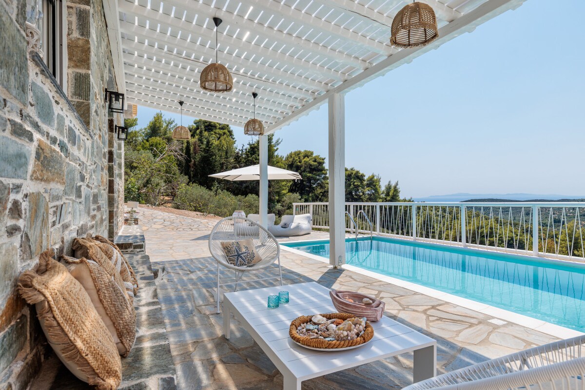 An outdoor lounge area features a spacious white table accompanied by comfortable seating, with light-colored cushions adding softness. A sparkling pool glistens under the sun nearby, while the expansive patio area is shaded by a white pergola, enhancing the relaxing atmosphere.