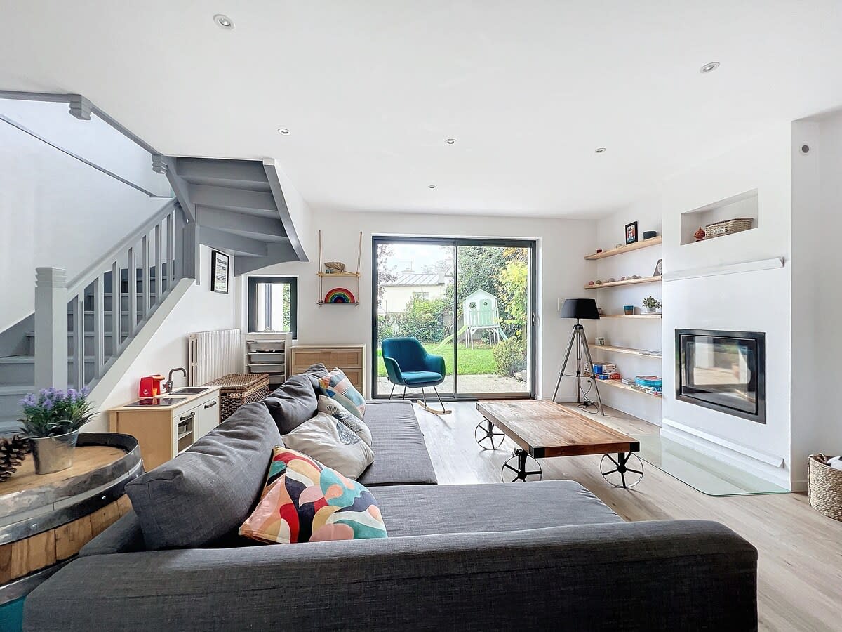 A spacious living area boasts a large sectional sofa with colorful cushions. An inviting coffee table is centered in front of the sofa. A sliding glass door opens to a garden view, enhancing natural light, while shelves display decorative items.