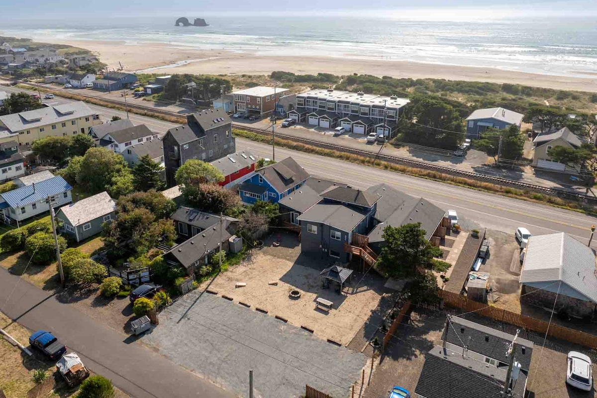 An aerial view captures a cluster of recently renovated cottages surrounded by greenery, with a pathway leading to the beach visible in the distance. The shoreline stretches along the horizon, while the spacious area in front offers a shared seating zone and access to nearby roads.