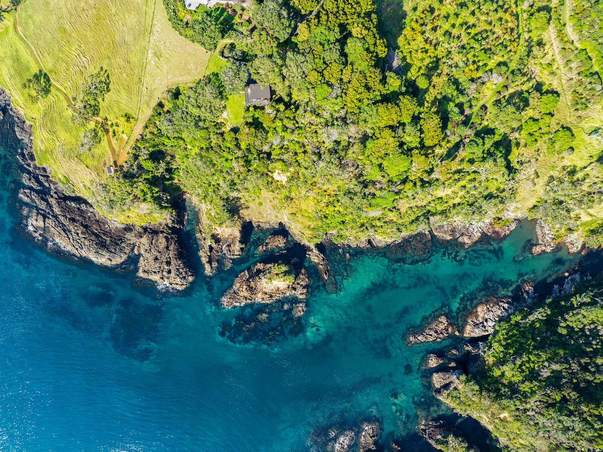 An aerial view of the coastal landscape shows jagged rocks emerging from the vibrant turquoise water. Lush greenery covers the land, interspersed with patches of open space and a glimpse of the Coastal Retreat tucked among the trees.