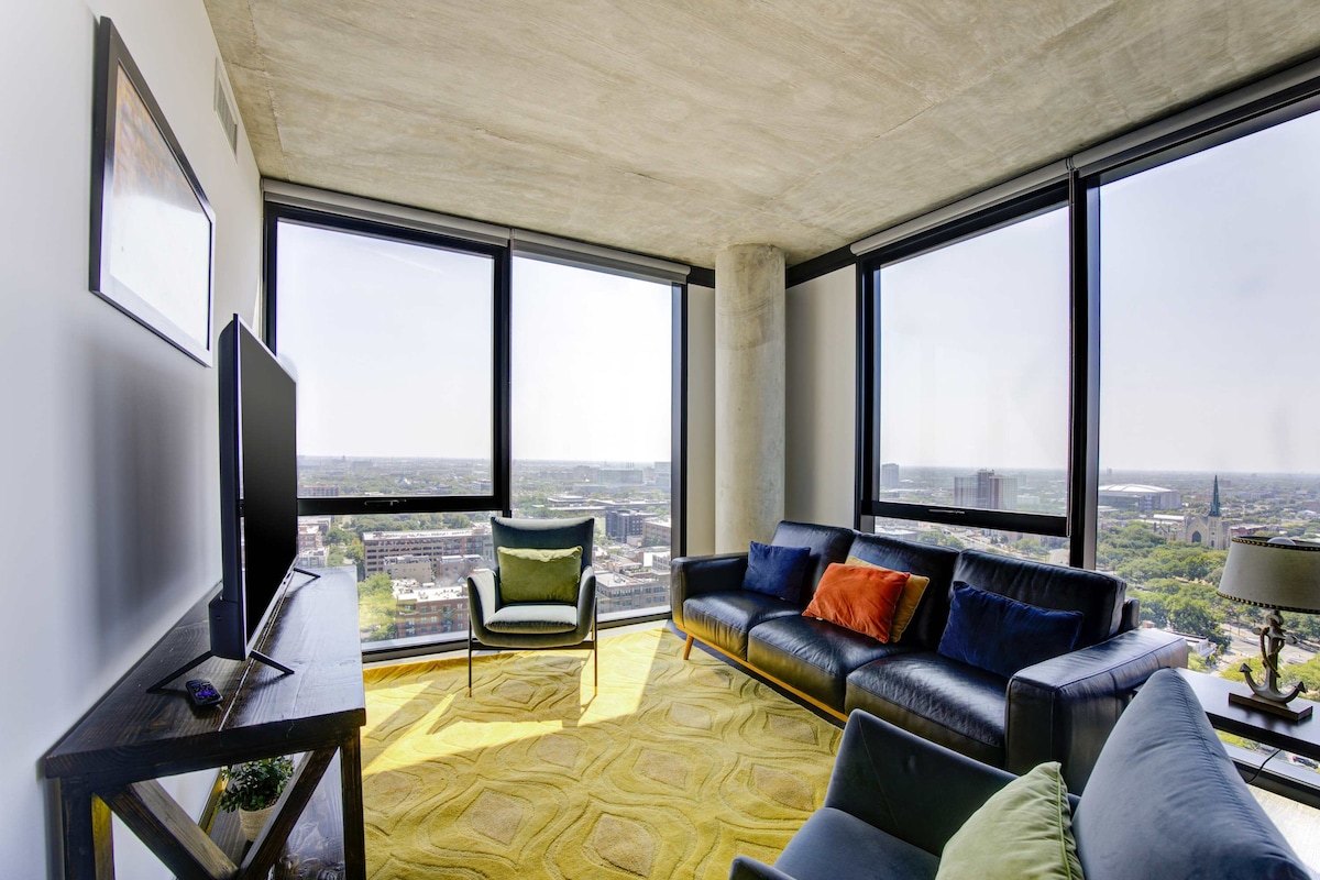 A modern living area features large windows that provide expansive views of the city. A sleek black sofa with orange and green accent pillows is positioned on a vibrant yellow area rug, complemented by a stylish chair in the corner. A flat-screen TV is mounted on the wall.