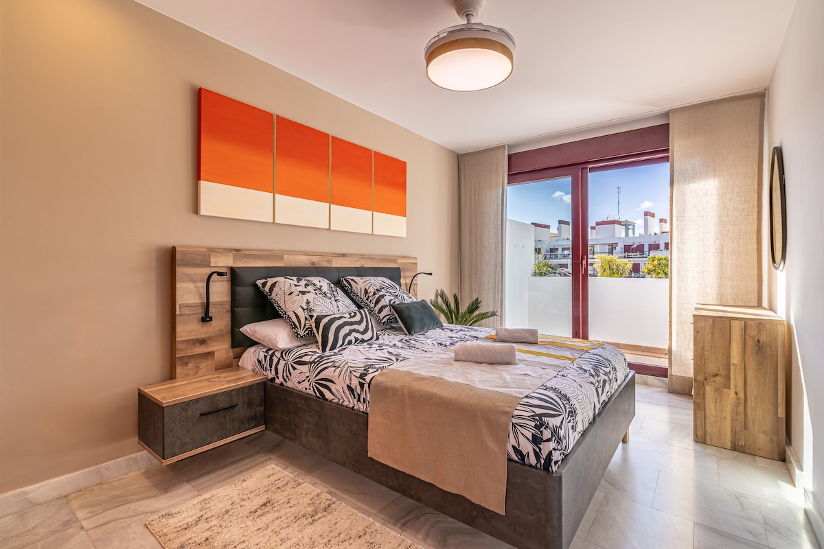 A spacious bedroom is presented with a large bed featuring a patterned duvet and soft towels. Two bedside lamps are situated on either side, and a wooden dresser is visible. Natural light streams in from the wide glass door leading to the terrace.