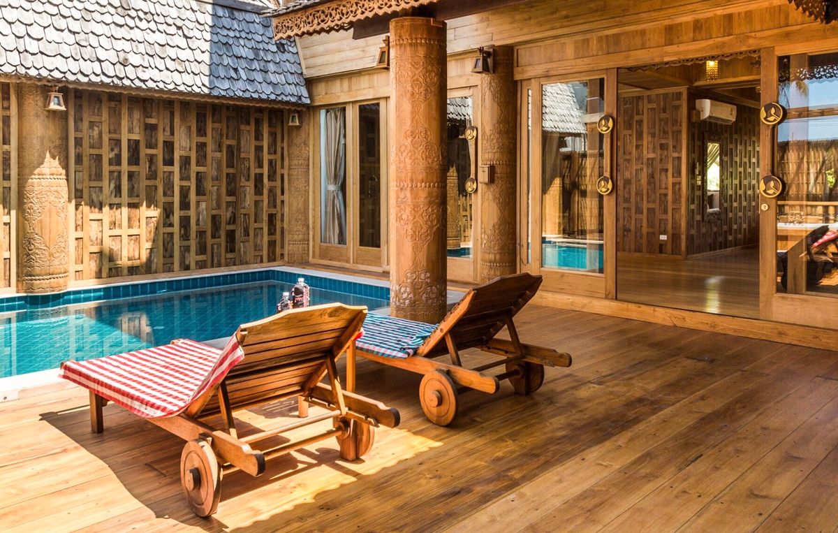 Two wooden sun loungers are positioned beside a private swimming pool. The loungers, crafted with natural materials, feature red and white striped fabric. Surrounding wooden decking and walls with intricate carvings enhance the tranquil setting, while large glass doors provide a view of the inviting interior.