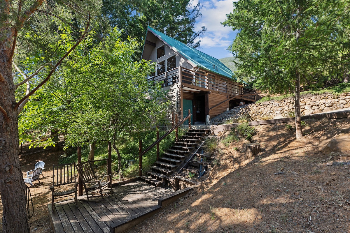 The exterior of the home is surrounded by trees, showcasing a sloped roof with green accents. A wooden staircase leads up to the entrance, while a seating area with wooden chairs is visible in the foreground, offering a peaceful retreat amidst the natural landscape.