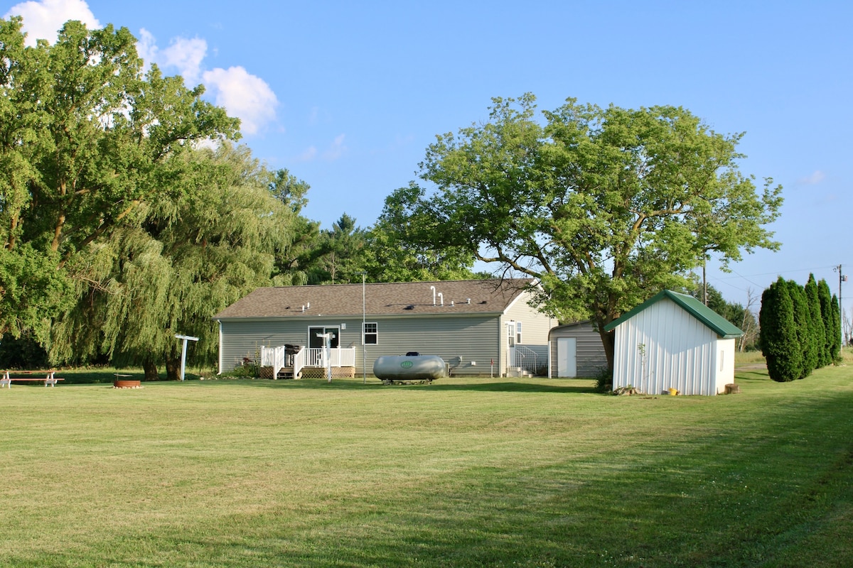 A single-story house is situated in a spacious yard, surrounded by lush greenery and trees. A shed is visible beside the home, and a canoe rests on the grass. Clear blue skies enhance the serene rural setting.