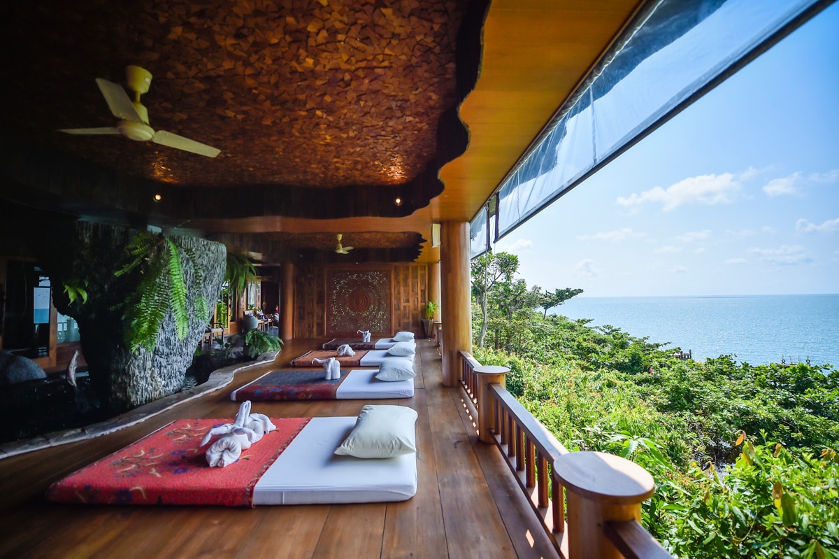 A spacious outdoor terrace features traditional Thai bedding arranged on polished wooden floors, overlooking the lush greenery and the sea. Large windows allow natural light to flood the area, enhancing the tranquil ambiance and providing an unobstructed view of the ocean beyond.