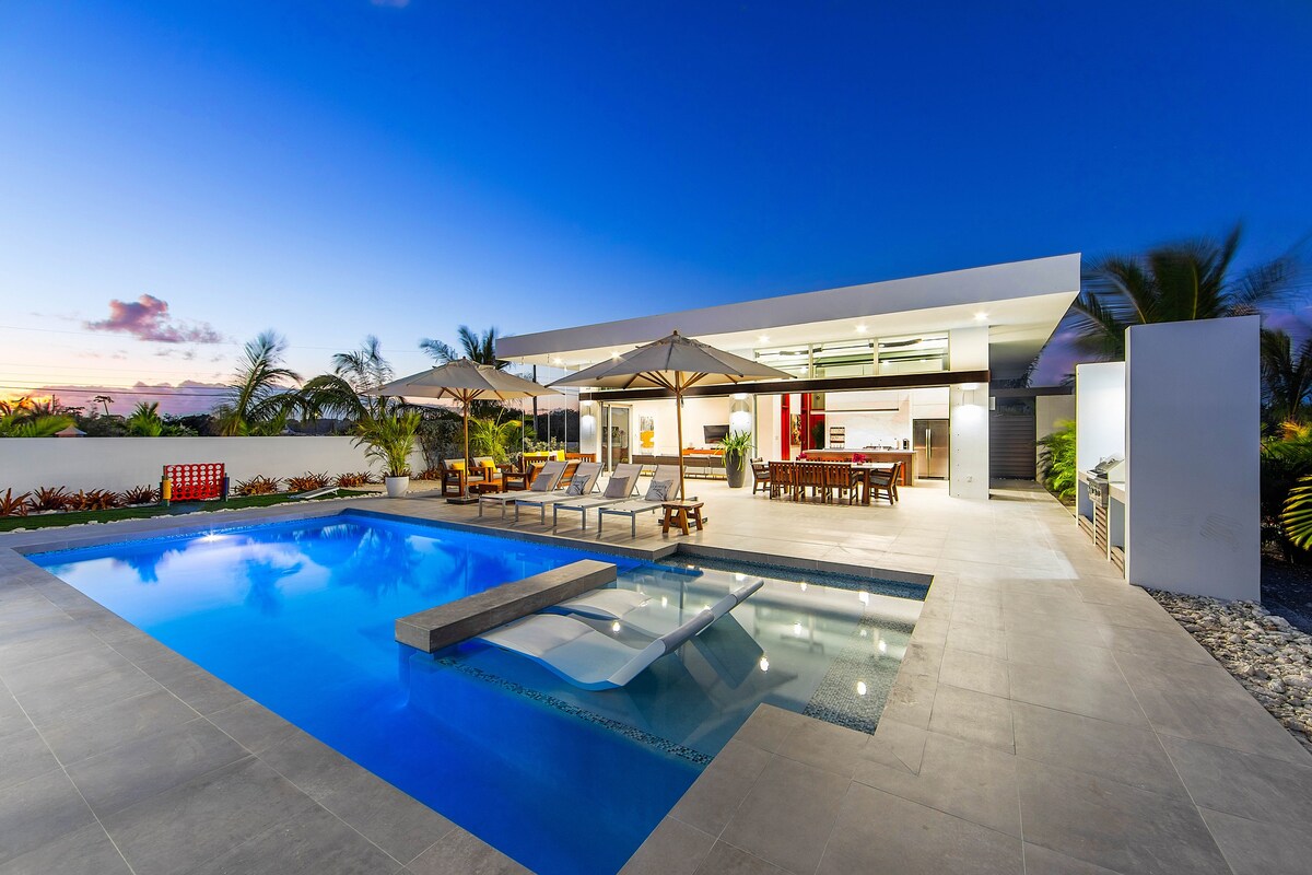 The outdoor area features a large private pool with a sunken lounge section, surrounded by elegant lounge chairs and shaded umbrellas. A spacious dining table is visible under the pavilion, providing an inviting space for gatherings. Lush landscaping complements the serene setting.