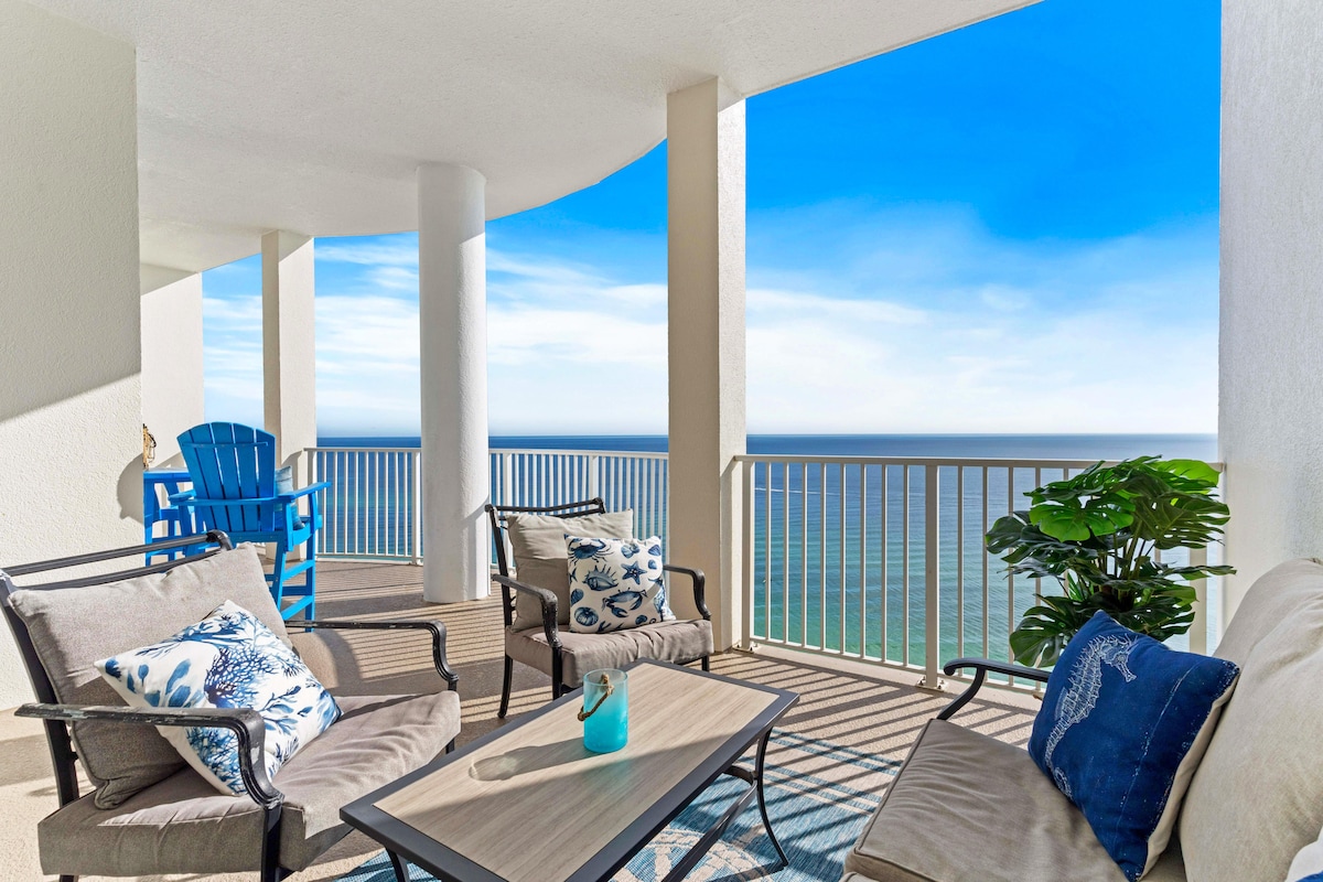 A spacious balcony features comfortable seating arrangements, including a pair of lounge chairs and a coffee table. The stunning ocean view is framed by a railing, while vibrant blue skies provide a picturesque backdrop. A potted plant adds a touch of greenery to the setting.