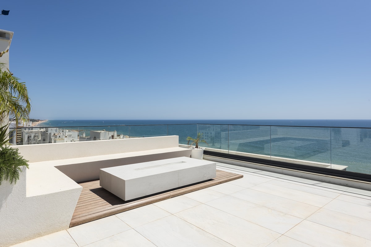 A spacious rooftop terrace features a large, minimalist lounge area with a stone platform and wooden decking. Glass railings provide unobstructed views of the ocean, while the bright sky enhances the serene atmosphere. Lush greenery adorns the space, adding a touch of nature.