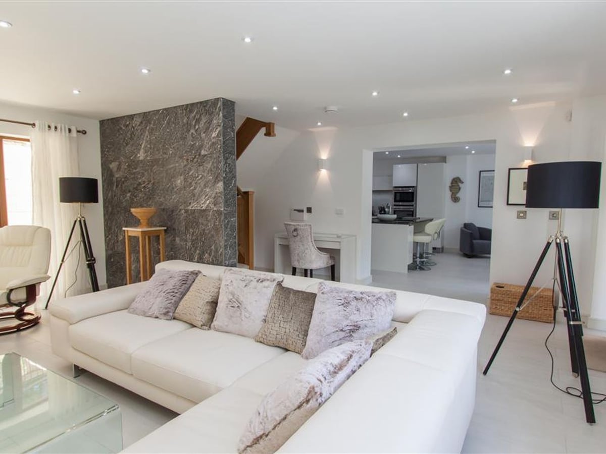 An open-plan living area features a light-colored sofa with decorative cushions. Floor lamps stand elegantly beside the seating area, and a sleek coffee table sits in front. A smooth transition to the dining and kitchen space is visible, hinting at an integrated design with modern finishes.