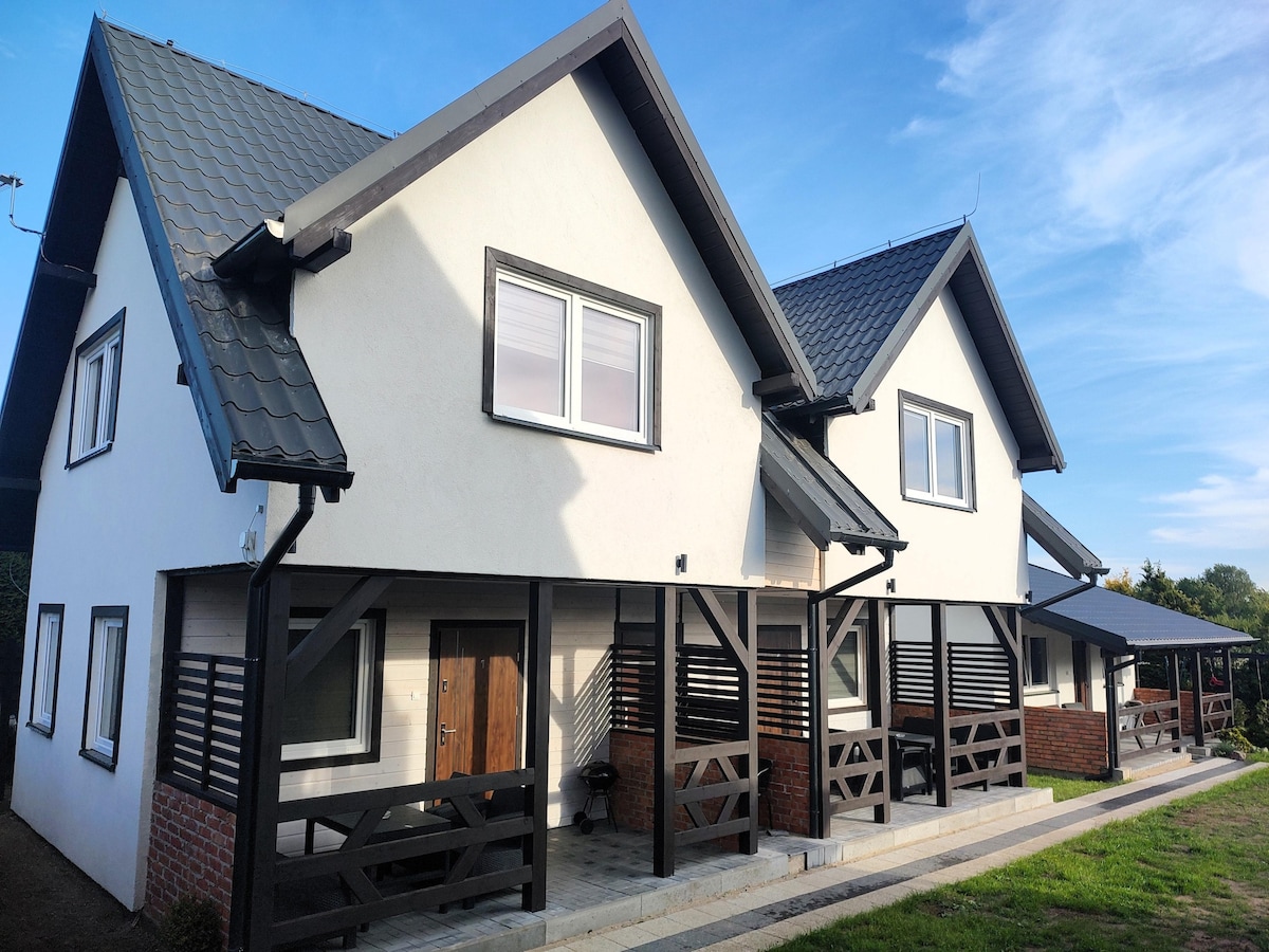 A two-story holiday home features a modern architectural design, highlighted by a pitched roof and a mix of white and dark exterior accents. Covered porches are visible, providing sheltered entryways. Large windows allow natural light to illuminate the interior spaces.