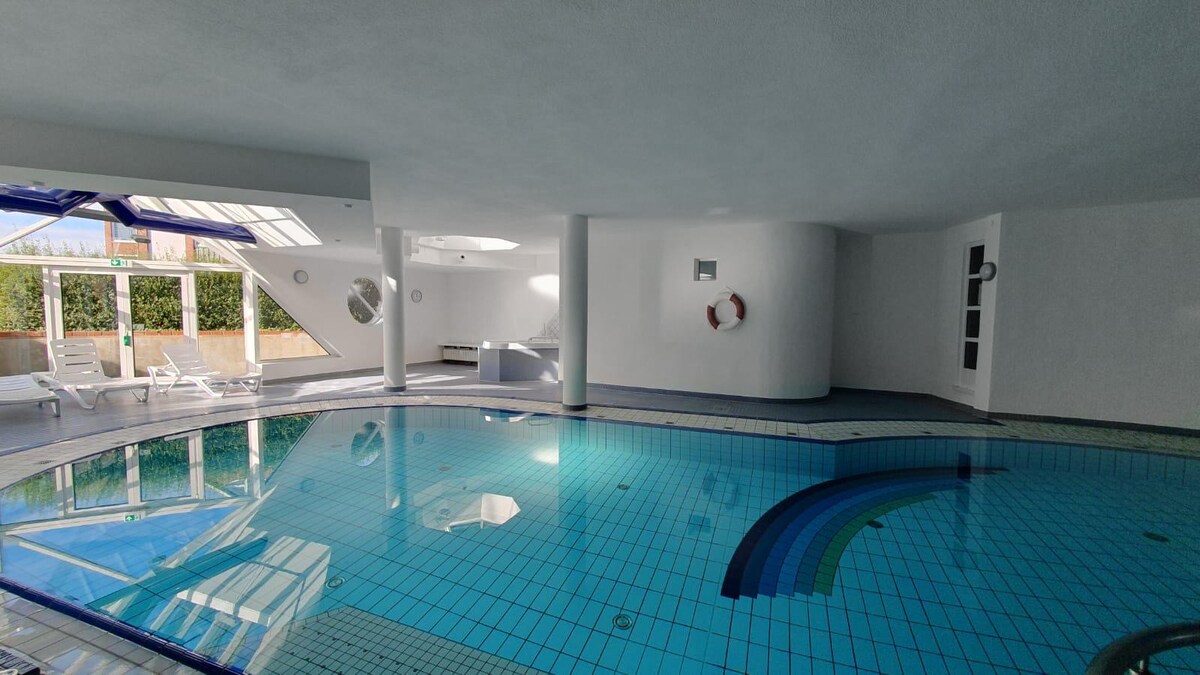 An indoor swimming pool is illuminated by natural light streaming through large windows. The pool features tiled walls and a tranquil water surface reflecting the space. Lounge chairs are arranged nearby, contributing to a relaxing atmosphere. Lifebuoys are mounted on the wall for safety.