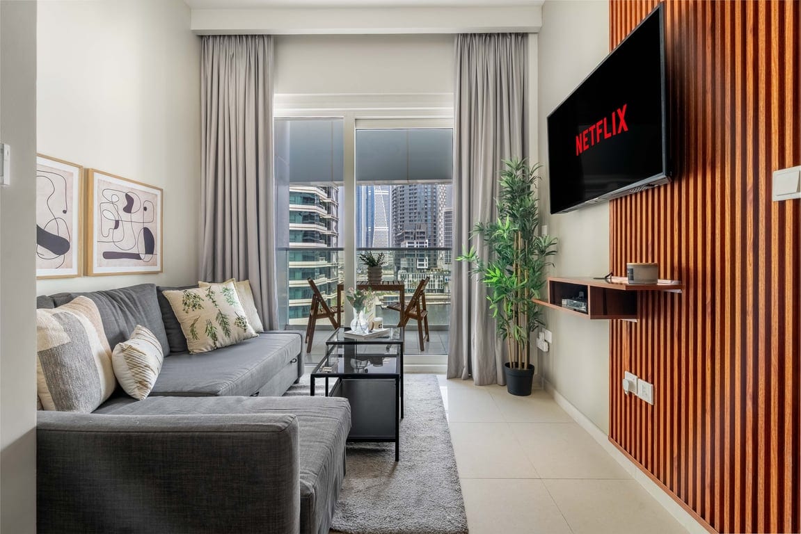 A modern living room features a plush grey sofa and a sleek coffee table, positioned in front of a wall-mounted TV displaying Netflix. Tall windows allow natural light to fill the space, with a small balcony visible outside, furnished with a dining table and chairs.