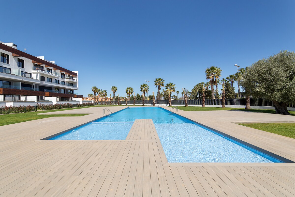 A rectangular swimming pool is framed by a wooden deck, surrounded by landscaped gardens and palm trees. The clear blue sky is reflected in the water, creating a serene and inviting outdoor space for relaxation.