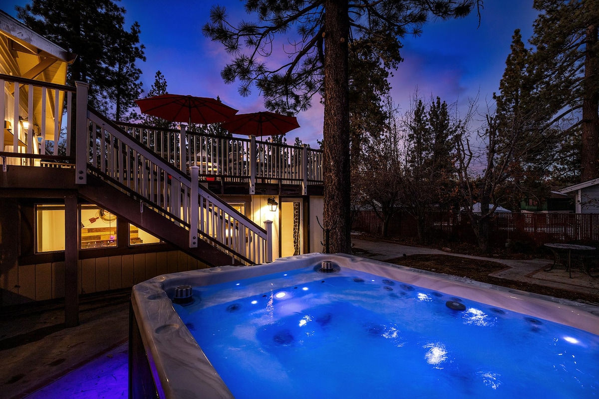 A hot tub is illuminated with blue lights, set against a serene evening sky. It is positioned on a stone patio, surrounded by trees. A staircase leads to an upper deck area with red umbrellas, providing a welcoming outdoor space.