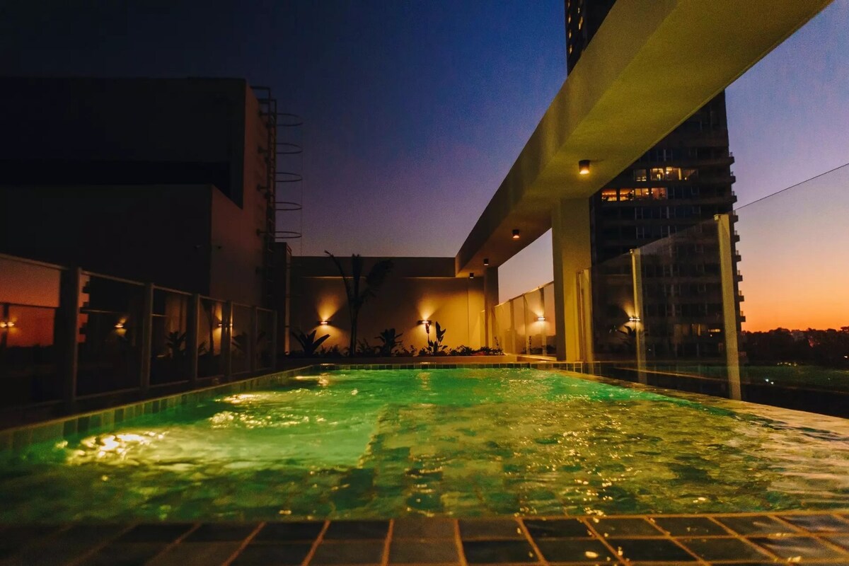 A rooftop pool is featured, with water illuminated in soft green hues against the twilight sky. The surrounding area includes clear glass railings and hints of palm plants, while the skyline forms a backdrop, creating a serene and spacious atmosphere.