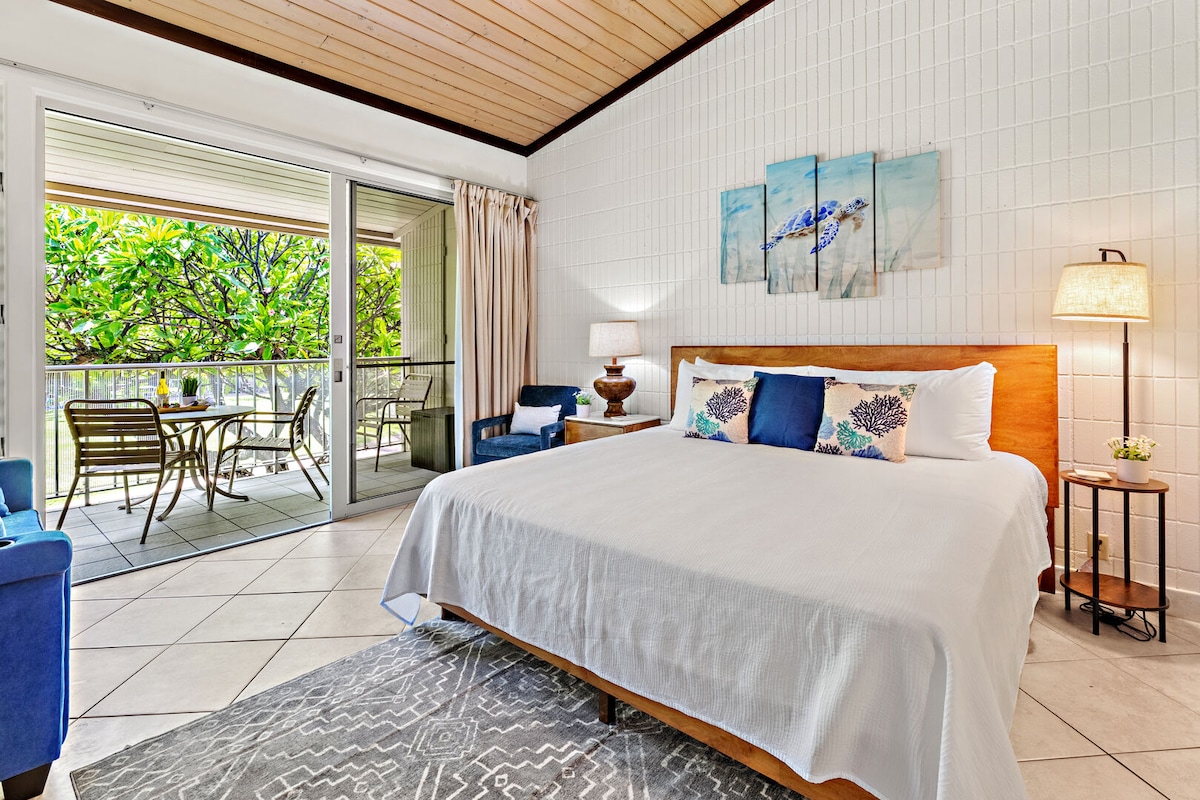A spacious studio condo featuring a plush king-size bed with decorative pillows. A subtle rug covers the tile floor. The room includes a seating area with blue chairs and a lamp. Sliding glass doors lead to a large lanai with outdoor seating amidst lush greenery.
