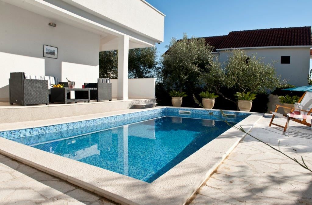 A private pool is framed by a clean patio area, featuring comfortable outdoor furniture in muted tones. Lush greenery surrounds the pool, complemented by potted plants. The clear blue water reflects the sunlight, enhancing the serene atmosphere.