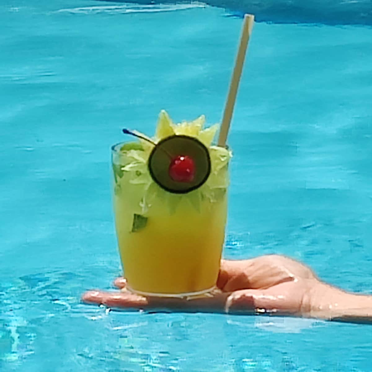 A refreshing cocktail is held above the surface of a clear blue pool. The drink is garnished with a lime slice, a maraschino cherry, and a decorative green garnish. Sunlight reflects off the water, creating a vibrant and inviting scene.