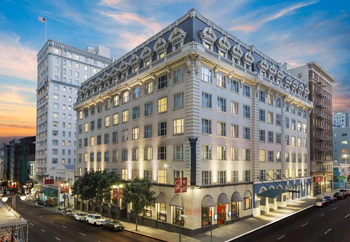 The exterior view of The Marker showcases its Beaux-Arts architecture with elegant detailing. Multiple stories feature large windows, illuminated at dusk, creating a welcoming ambiance. The building is framed by trees and streetlights, set against a softly lit sky.