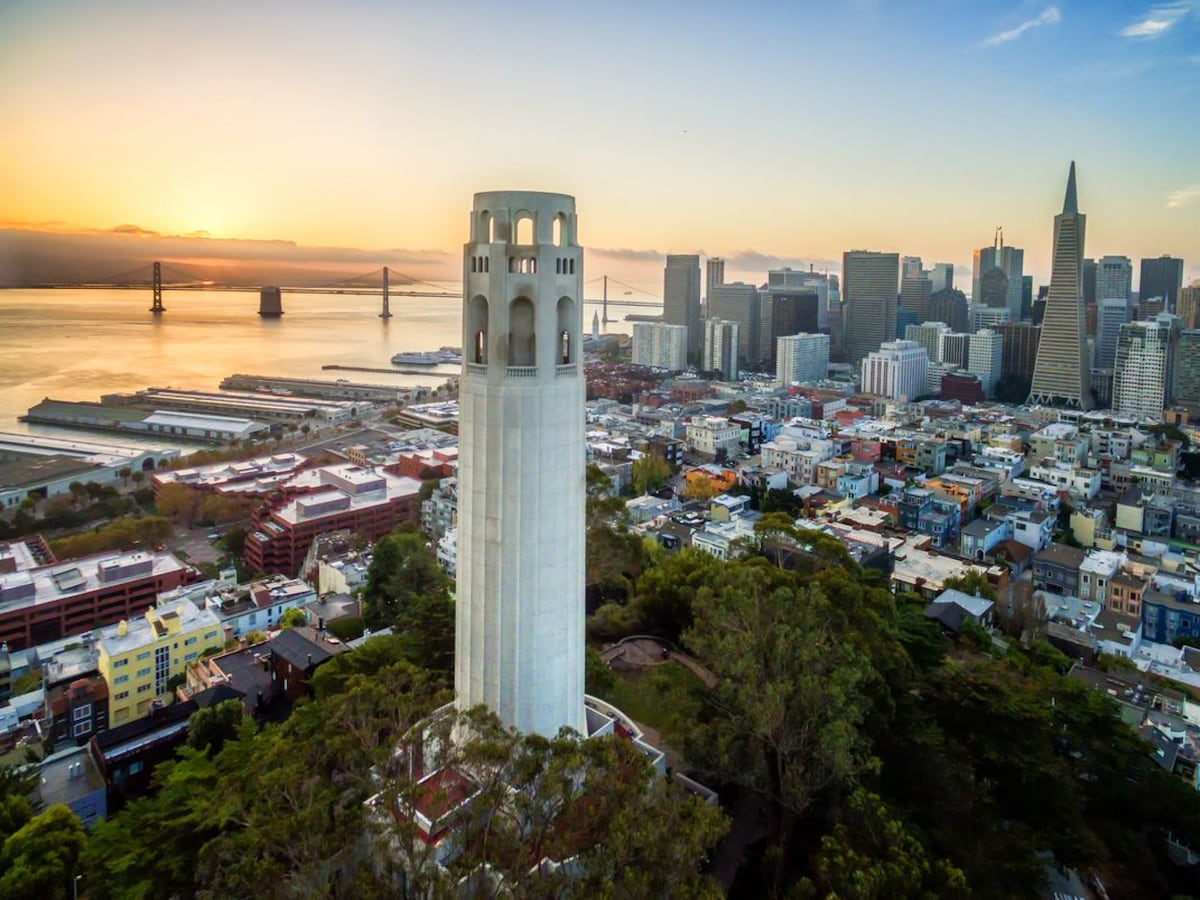 An aerial view captures San Francisco's iconic Coit Tower, standing prominently amidst the landscape. The scene is enriched by a golden sunset, illuminating the skyline and the Bay Area. A blend of urban architecture and natural greenery surrounds the tower, offering a sense of tranquility.