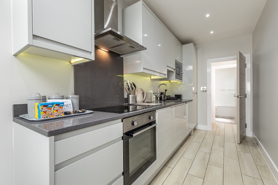 A modern kitchen is shown, featuring sleek, high-gloss cabinetry and a dark countertop. Top-of-the-line appliances, including an oven, microwave, and induction cooktop, are present. A sink and various kitchen utensils are organized neatly. A door in the background leads to another space.