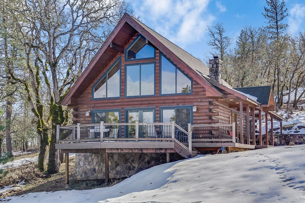 The spacious log cabin features a large deck overlooking a snowy landscape. Expansive windows allow natural light to fill the interior while providing views of the surrounding trees. The sloped roof adds to the rustic charm of the architecture.