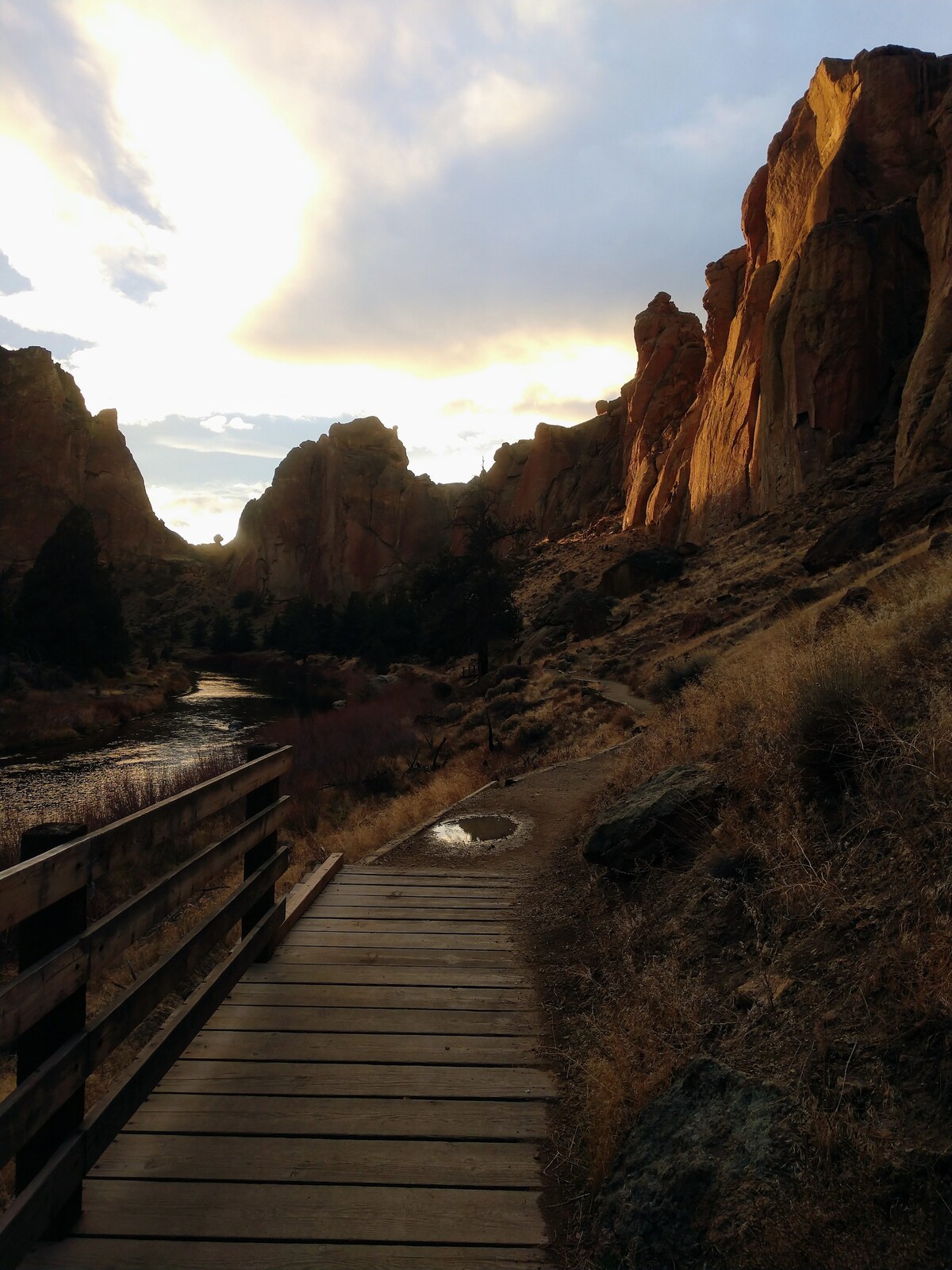 A wooden pathway winds through a natural landscape, bordered by rugged rock formations and a flowing river. Soft, golden sunlight bathes the cliffs, while trees provide green accents. The scene conveys a sense of tranquility in the wilderness.