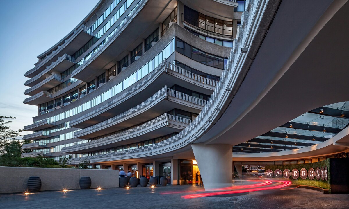 The exterior of the Watergate Hotel is showcased, highlighting the flowing architecture with rounded balconies and expansive glass windows. The entrance is illuminated, revealing the hotel's signage, while modern landscaping and smooth paths create a welcoming approach. The evening light enhances the building's contemporary design.