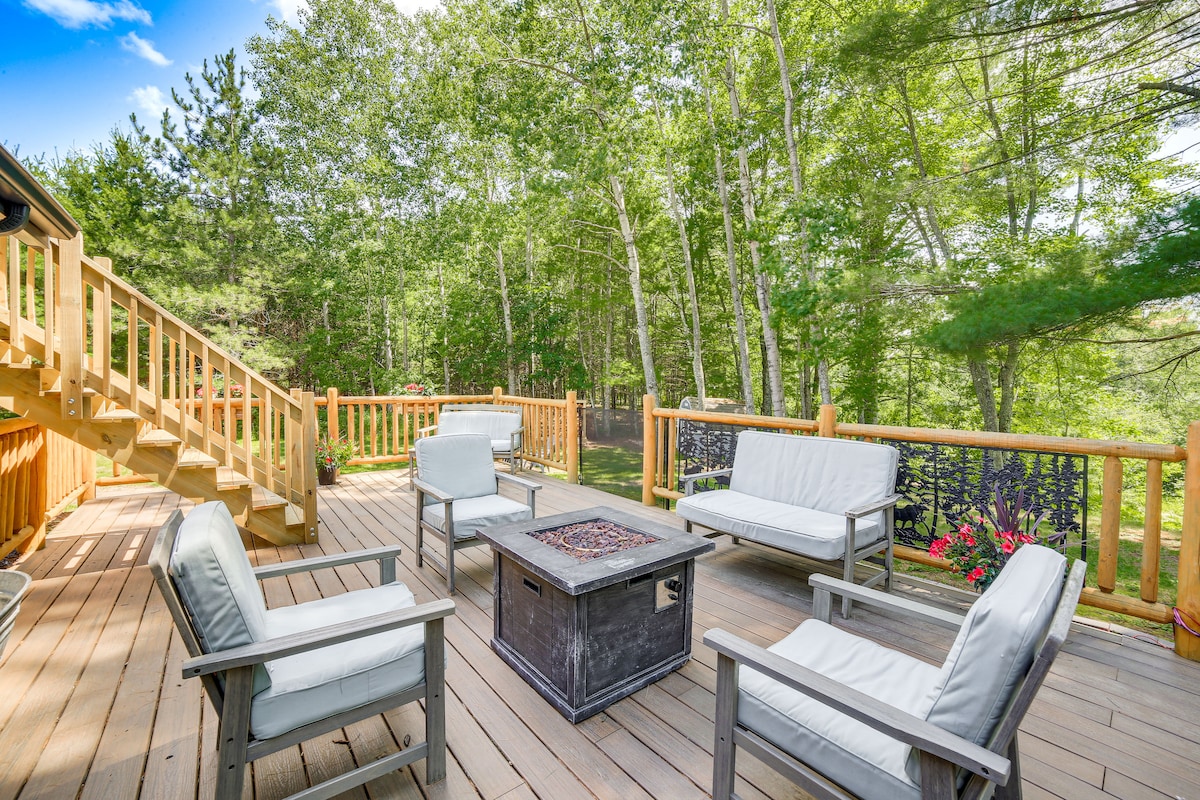 A spacious 2-story deck is featured, furnished with four white armchairs surrounding a central fire table. Lush greenery from surrounding trees creates a natural backdrop, contributing to a serene outdoor ambiance. A spiral staircase leads to the upper level of the cabin.