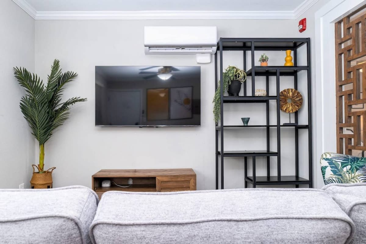 A living area is featured, showcasing a mounted flat-screen TV on the wall. A wooden TV stand is positioned below, with a black shelving unit beside it displaying decorative items and plants. A plush gray sofa is visible in the foreground, alongside a leafy indoor plant.