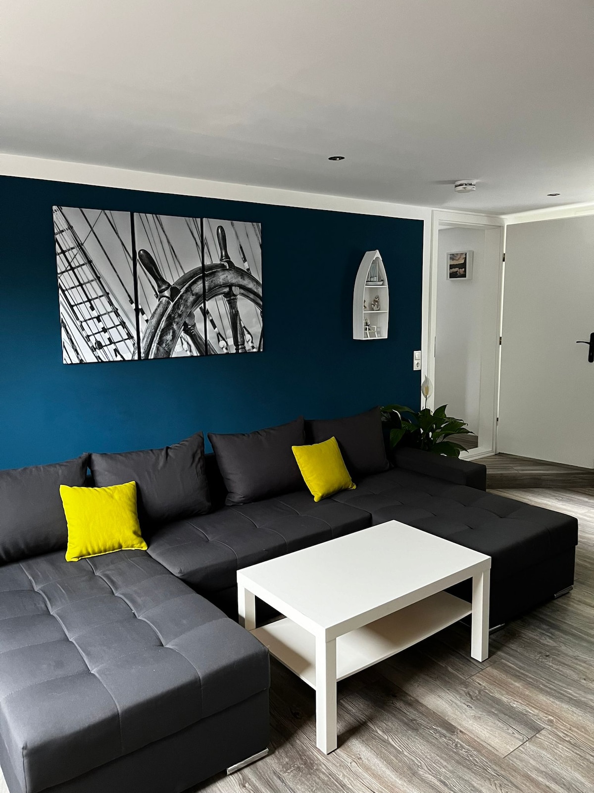 A modern living area features a dark sectional sofa adorned with bright yellow cushions. A white coffee table sits in the center, complemented by a dark blue accent wall with nautical-themed artwork above. A small plant adds a touch of greenery to the space.