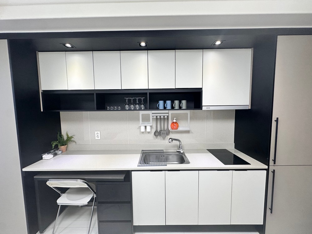 A modern kitchen area features sleek black and white cabinetry with under-cabinet lighting. Essential appliances, including a stainless steel sink and cooktop, are easily accessible. Organized kitchenware is displayed on the wall rack, complementing the minimalist design.