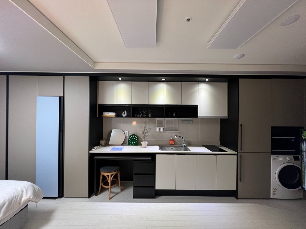 A compact kitchenette features modern cabinetry in neutral tones, complemented by a small dining table with a wooden stool. Appliances include a refrigerator, a washing machine, and a sink, with a window providing natural light to the space.